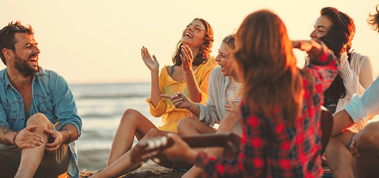 Happy friends sitting on the beach singing and playing guitar during the sunset