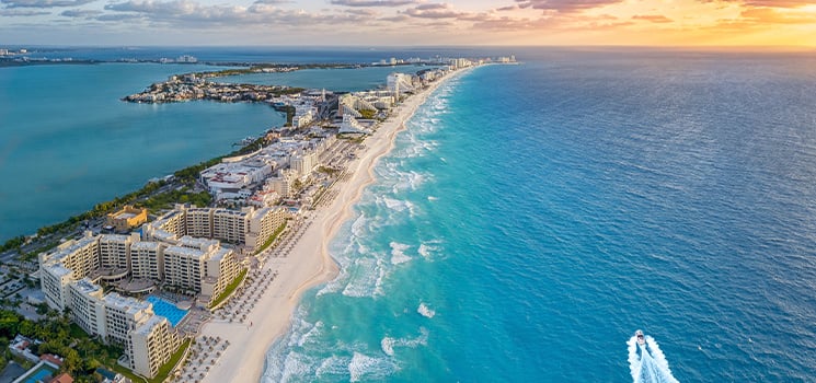 Panoramic aerial view of Cancun's hotel zone with pristine beaches, turquoise Caribbean Sea, and a boat cruising along the coastline at sunset.
