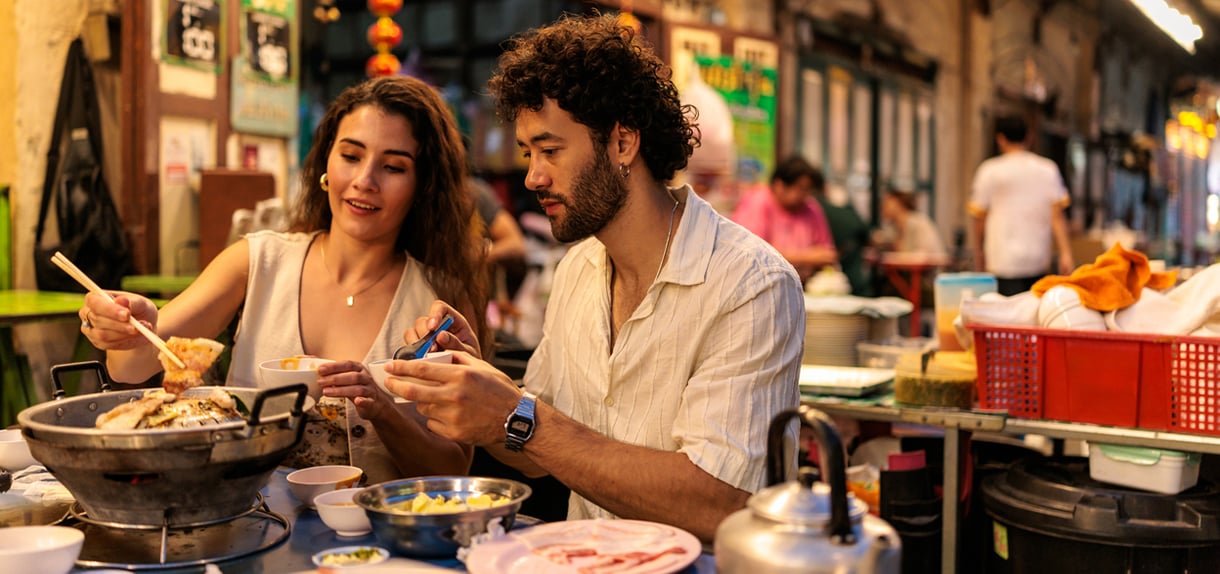 Couple enjoying street food at bustling Thai market stall with colorful dishes and vendors.