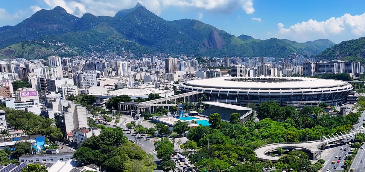 Aerial view of Maracanã Stadium in Rio de Janeiro, surrounded by dense city buildings and green mountains under a blue sky.