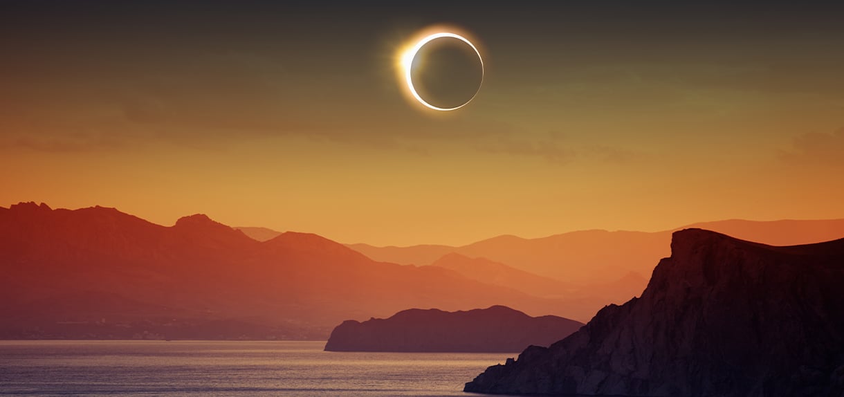 Solar eclipse with bright corona visible over mountainous coastline landscape in golden twilight sky.