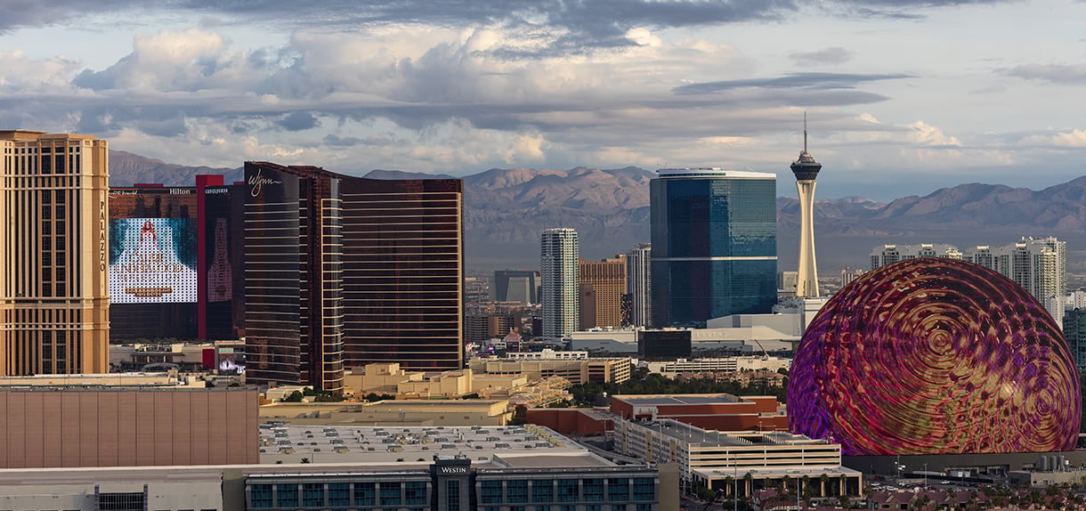 Aerial view of the Las Vegas Strip skyline with the Sphere venue displaying a colorful pattern, the Stratosphere tower, and desert mountains in the background.