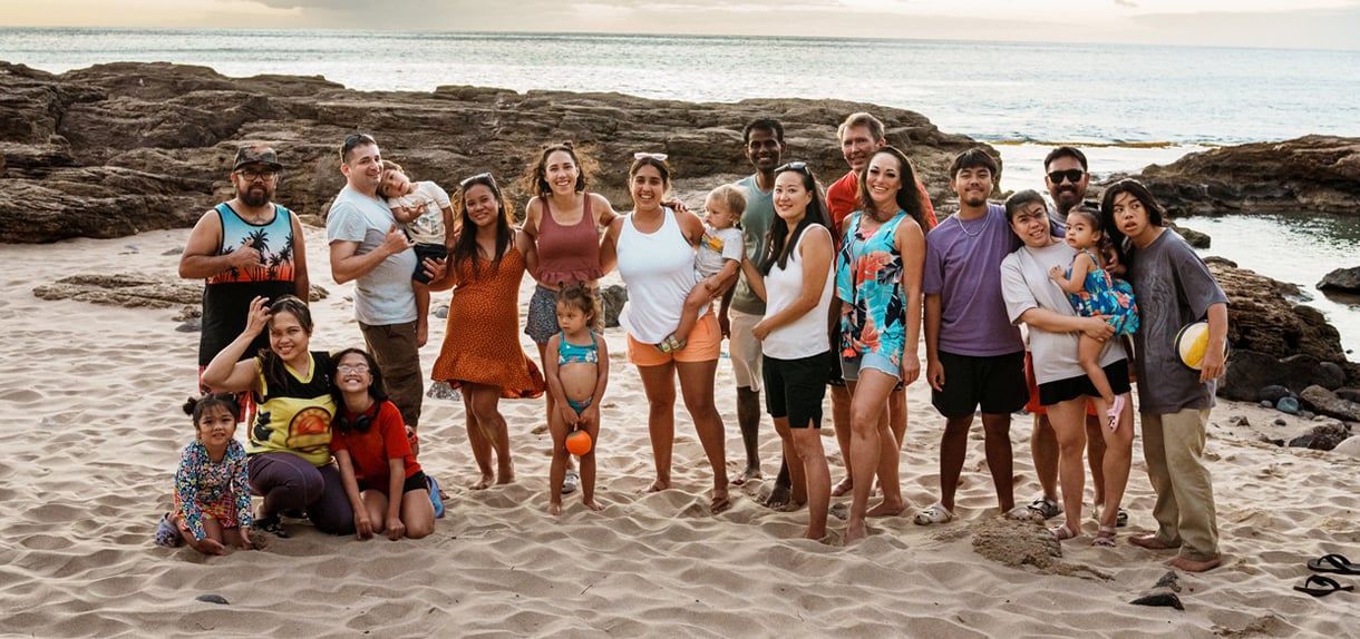 Extended family of multiple generations poses together on sandy beach with rocky coastline and ocean backdrop.