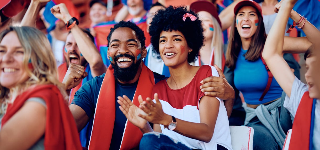 A diverse group of enthusiastic sports fans celebrating in stadium seats, wearing team colors and face paint while cheering.