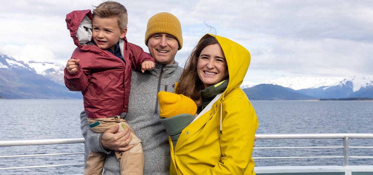 Happy family of three in winter gear smiling on cruise ship deck with Alaskan mountains behind.