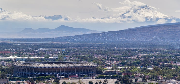Aerial view of Estadio Azteca in Mexico City surrounded by suburban neighborhoods with snow-capped mountains in background.